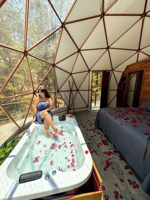 Une tente confortable avec un jacuzzi et un lit, entourée d’une forêt.