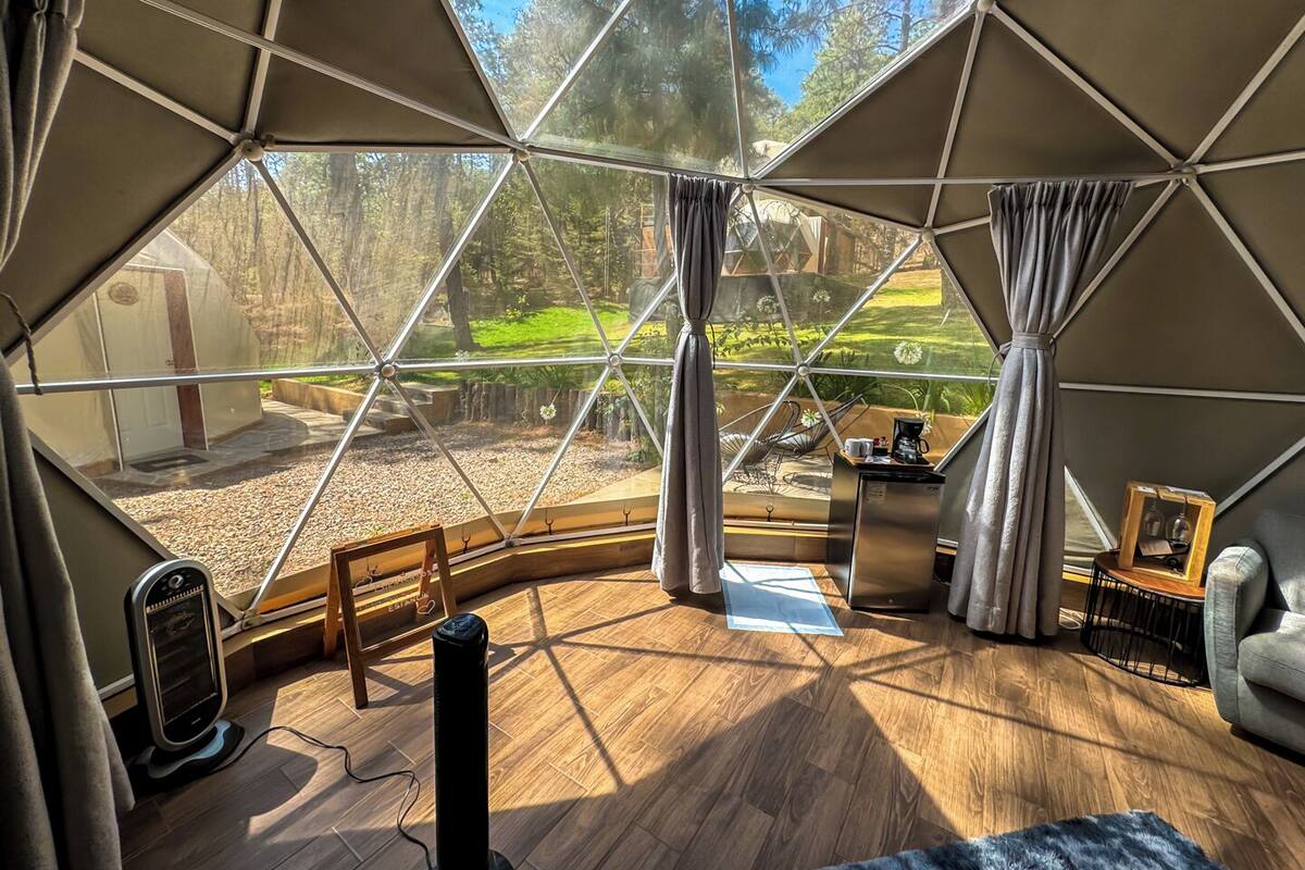 Ein Raum in Form einer Geodätischen Kuppel mit Holzboden, einem kleinen Tisch mit Kaffeemaschine und Blick auf einen Garten durch große Fenster.