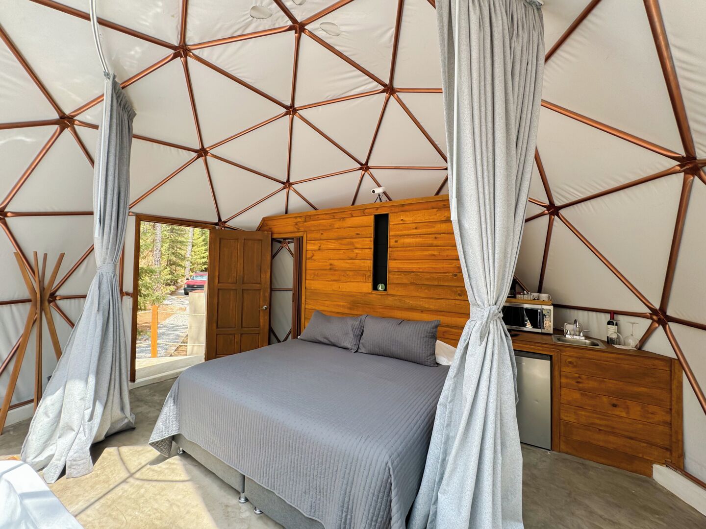 A bedroom with a bed, curtains, a wooden headboard, a small wooden cabinet, and a window.