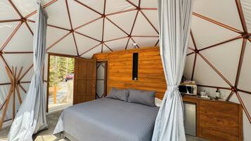 A bedroom with a bed, curtains, a wooden headboard, a small wooden cabinet, and a window.