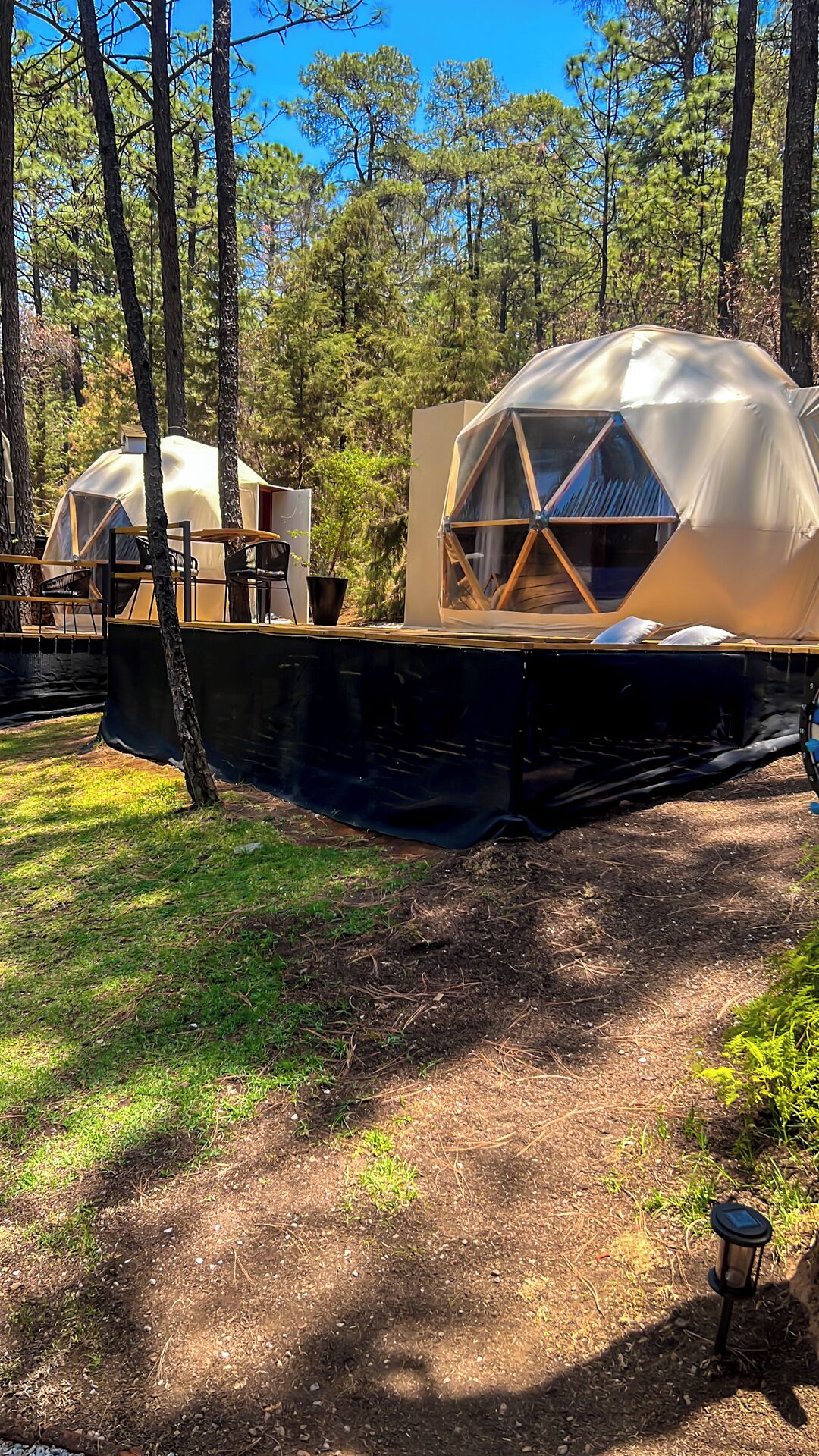 Two dome-shaped tents with a wooden frame, set in a forested area with a dirt ground.