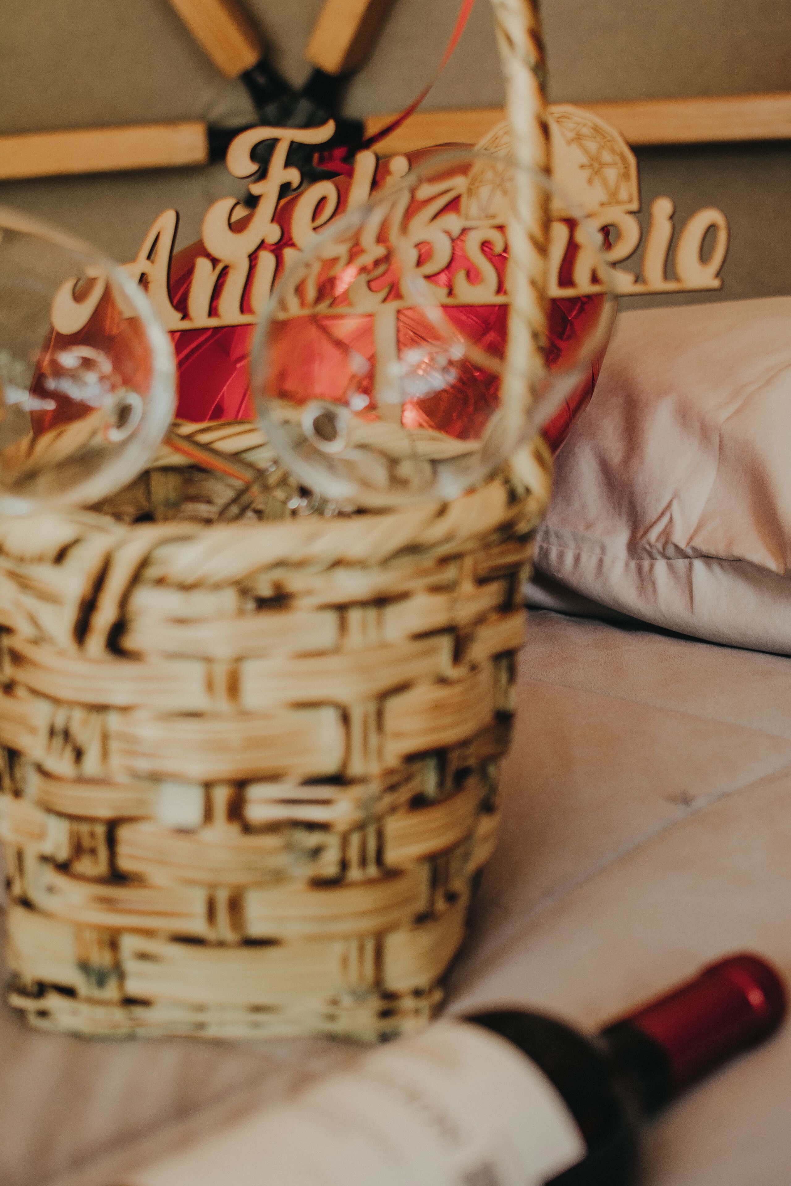 A wicker basket with a red ribbon, containing a bottle and a decorative item with the text "Feliz Aniversario".