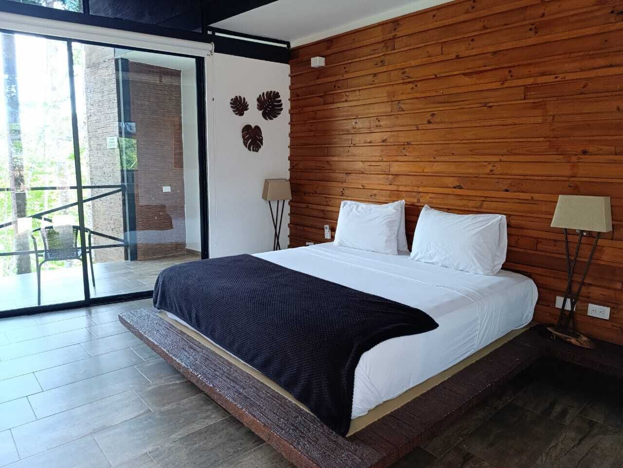 A bedroom with a large bed, wooden panel walls, a glass door leading to an outdoor area, and a nightstand with a lamp.