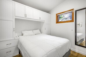 A bedroom with a large bed, white cabinets, a window with a view, and a mirror. - Cozy Cottage by the Lakeshore #4 (West Kelowna)