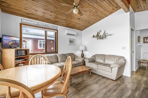 Ein gemütliches Wohnzimmer mit Holzbalkendecke, einem großen Fenster, einem Fernseher, einer Couch, einem Esstisch und einer Lampe.