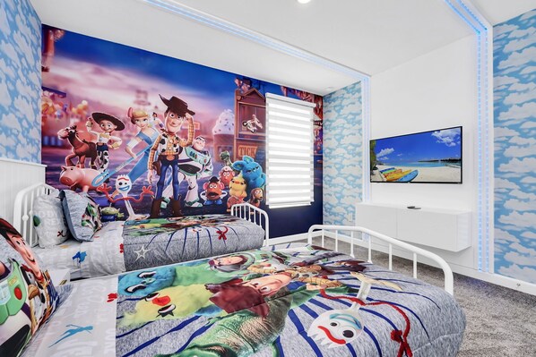 Een slaapkamer met twee bedden, een grote muurschildering van tekenfiguren, een televisie en een raam met jaloezieën.