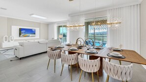 A modern dining area with a wooden table, white upholstered chairs, and a large window with a view of a pool. - Ultimate Bespoke 6 Bedroom Champions Gate Pool Villa (Davenport)