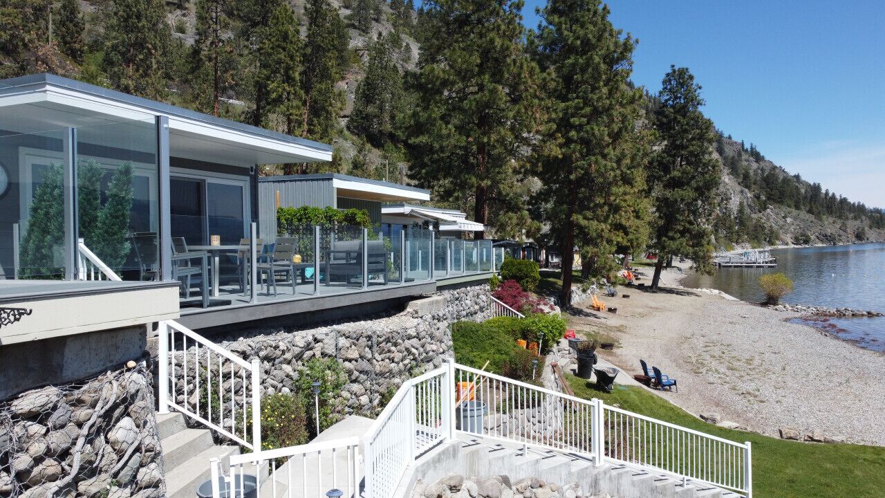 Ein modernes Haus mit großen Glasfenstern, einer Terrasse und Blick auf einen See und einen Wald.