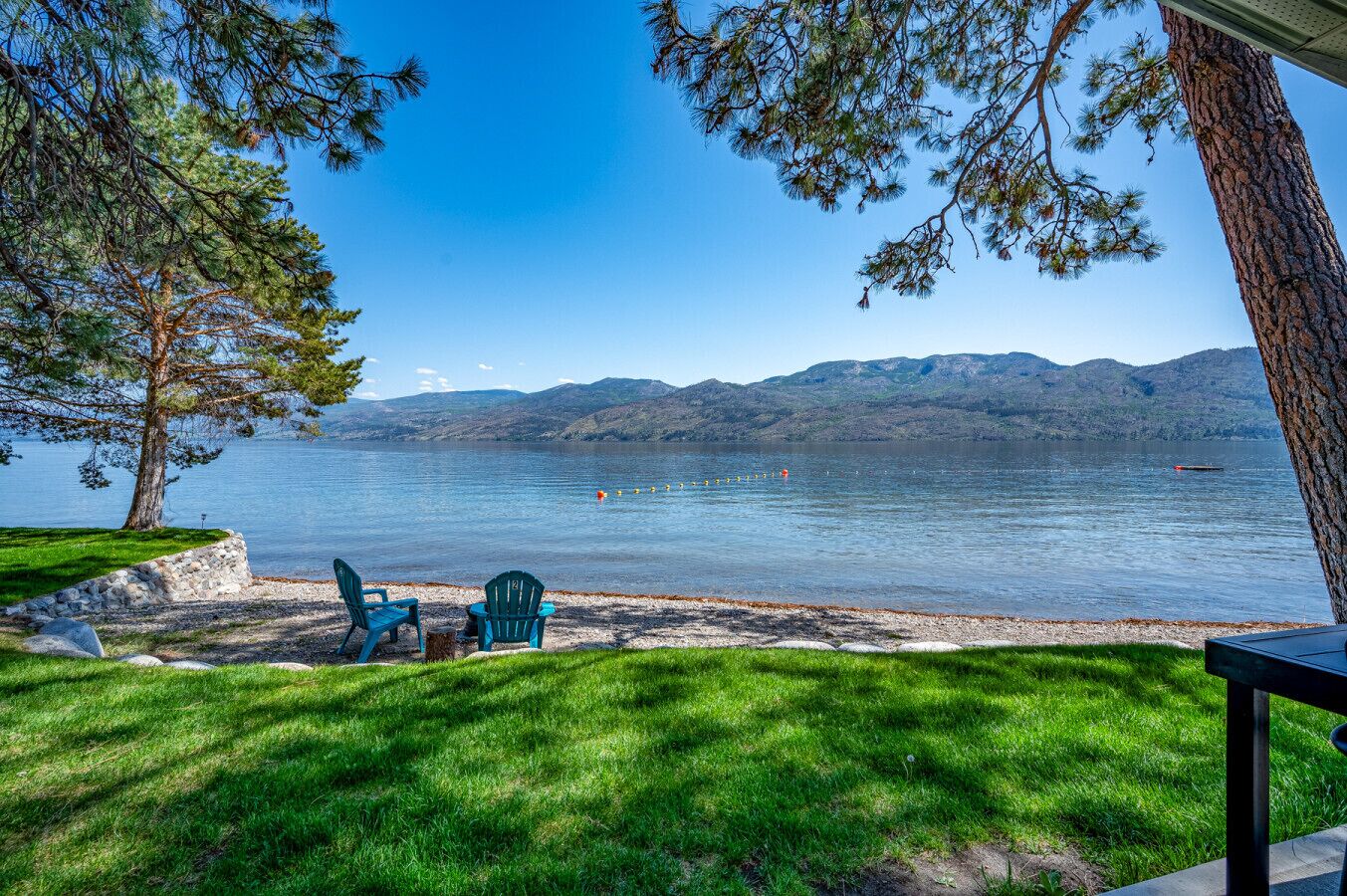 Vista al lago con un espacio cubierto de césped, dos sillas y una mesa bajo un árbol.