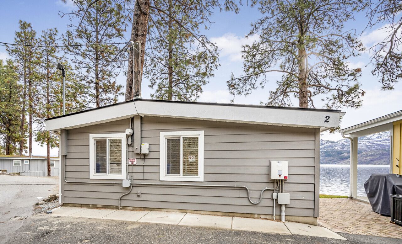 A small, single-story cabin with two windows, a covered outdoor seating area, and a view of a lake.