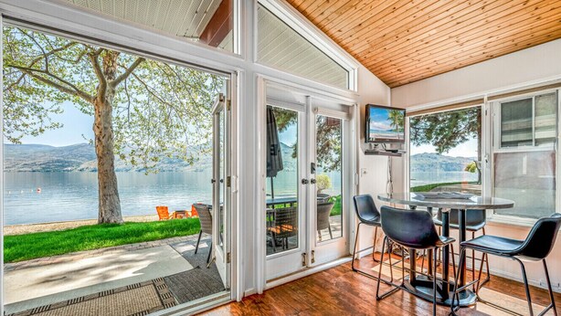 Een ruime kamer met een grote schuifglazen deur die leidt naar een terras met uitzicht op een meer en bergen. Binnen staat een ronde tafel met stoelen en is er een televisie aan de muur bevestigd.