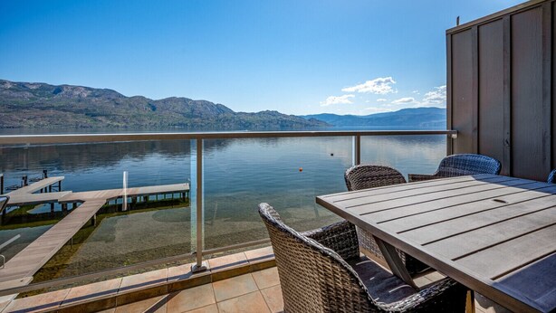 Varanda com mesa de madeira e cadeiras de vime, com vista para um lago calmo e montanhas.