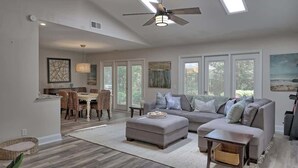 A spacious living room with a gray sofa, ottoman, and a dining area with wicker chairs.