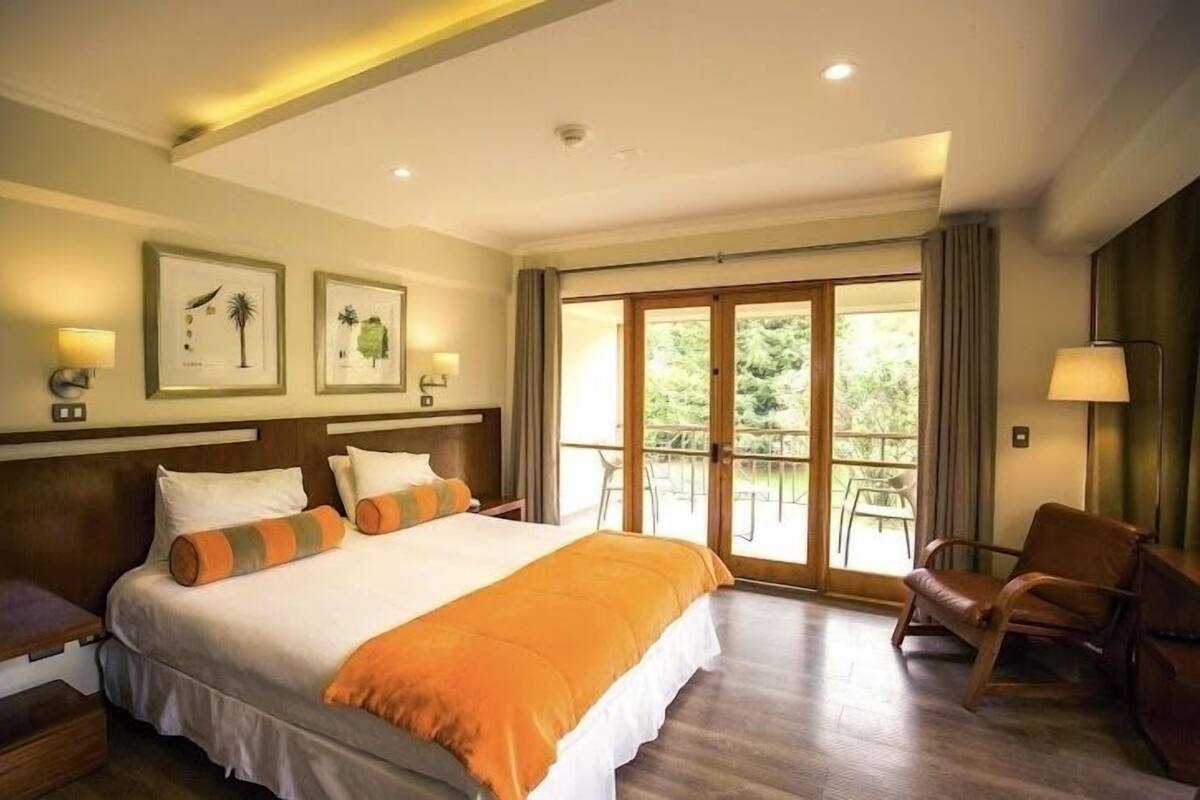Una habitación de hotel con una cama grande, cabecera de madera, un balcón con sillas y cuadros en la pared.