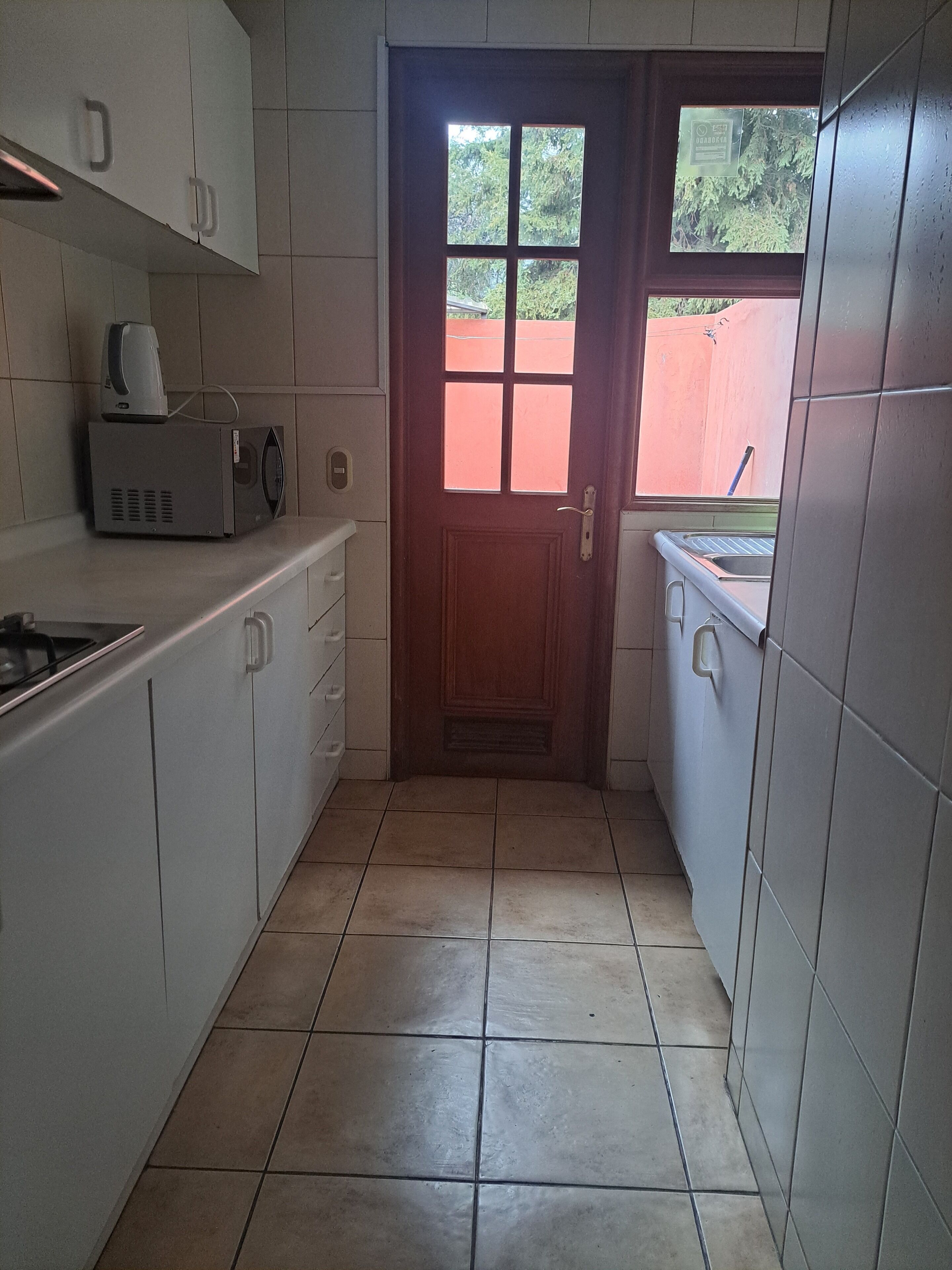 A narrow kitchen with white cabinets, a microwave, a sink, and a window with a view of greenery outside.
