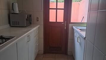 A narrow kitchen with white cabinets, a microwave, a sink, and a window with a view of greenery outside.