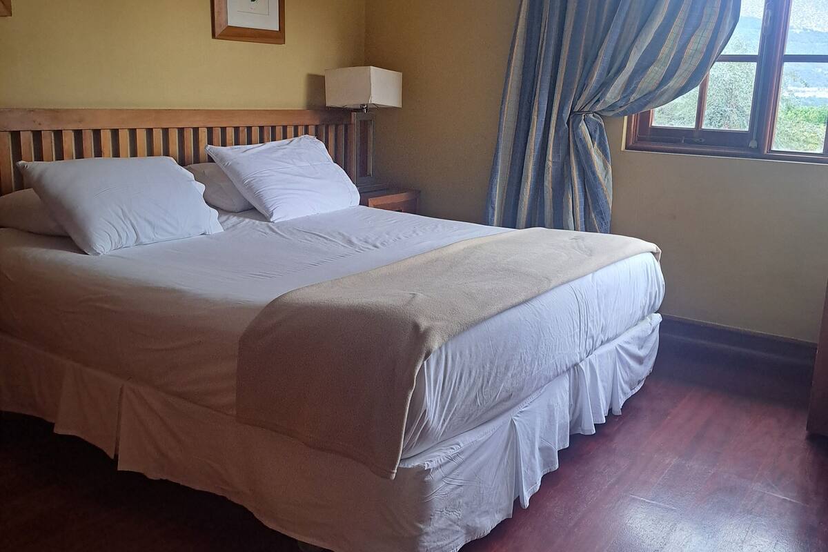 Una cama bien hecha con sábanas blancas en una habitación con suelo de madera, una mesita de noche con lámpara y una ventana con cortinas.