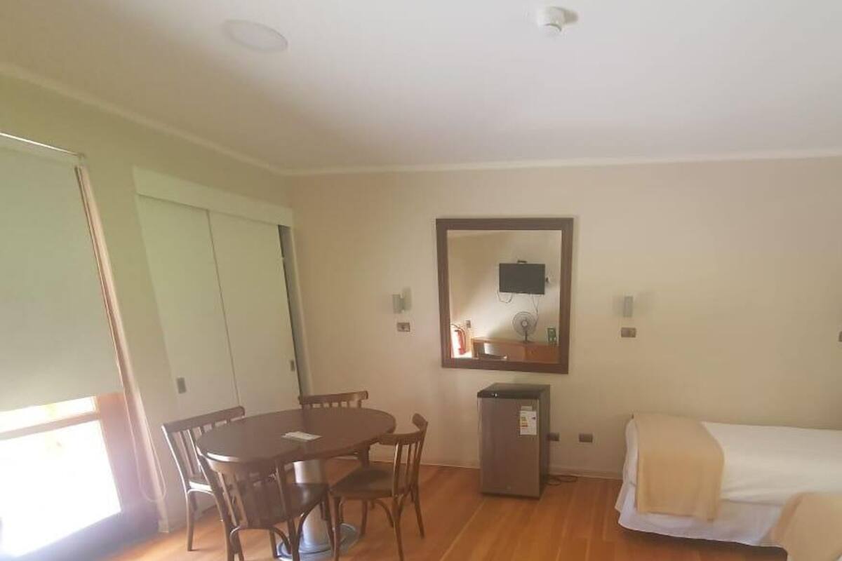 Una habitación con una cama, una pequeña mesa con sillas, un refrigerador y un espejo en la pared.