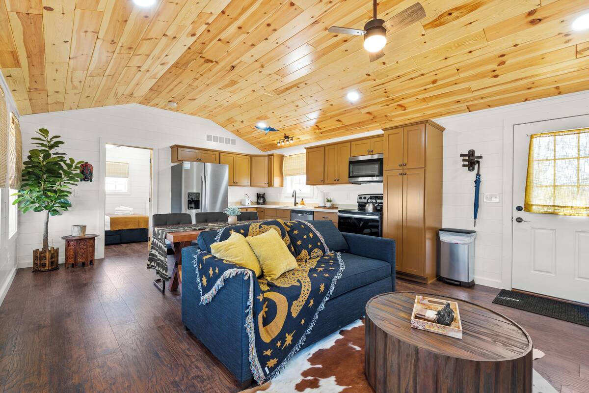 A spacious living room with a blue sofa, wooden ceiling, and a kitchen area with cabinets and appliances.