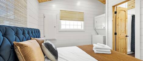 A bedroom with a tufted headboard, a large window with blinds, a white bed with a brown blanket, and a wooden door leading to a bathroom.