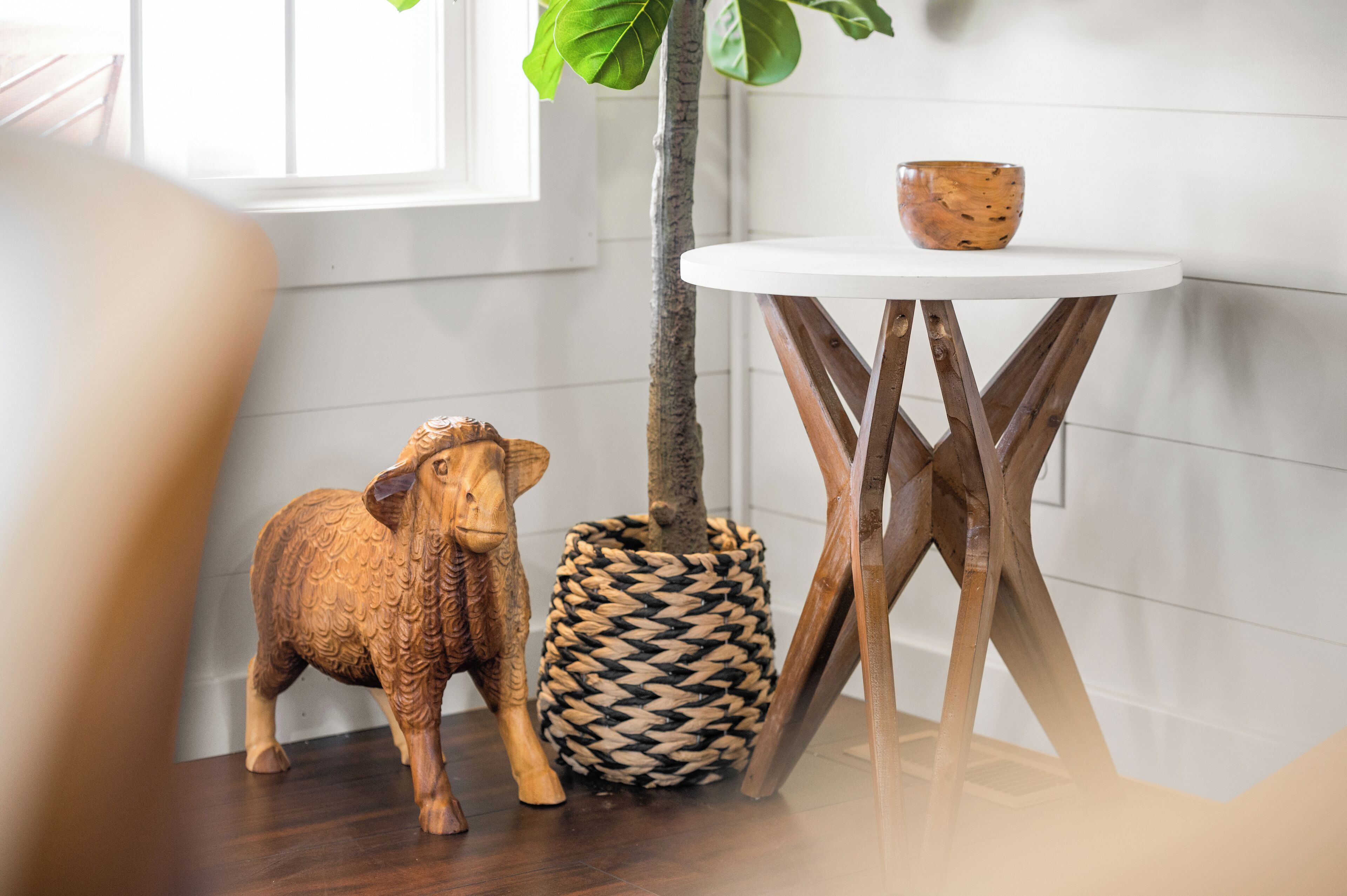 A small round table with a woven basket and a wooden bowl on top, a wooden sculpture, and a tree trunk.
