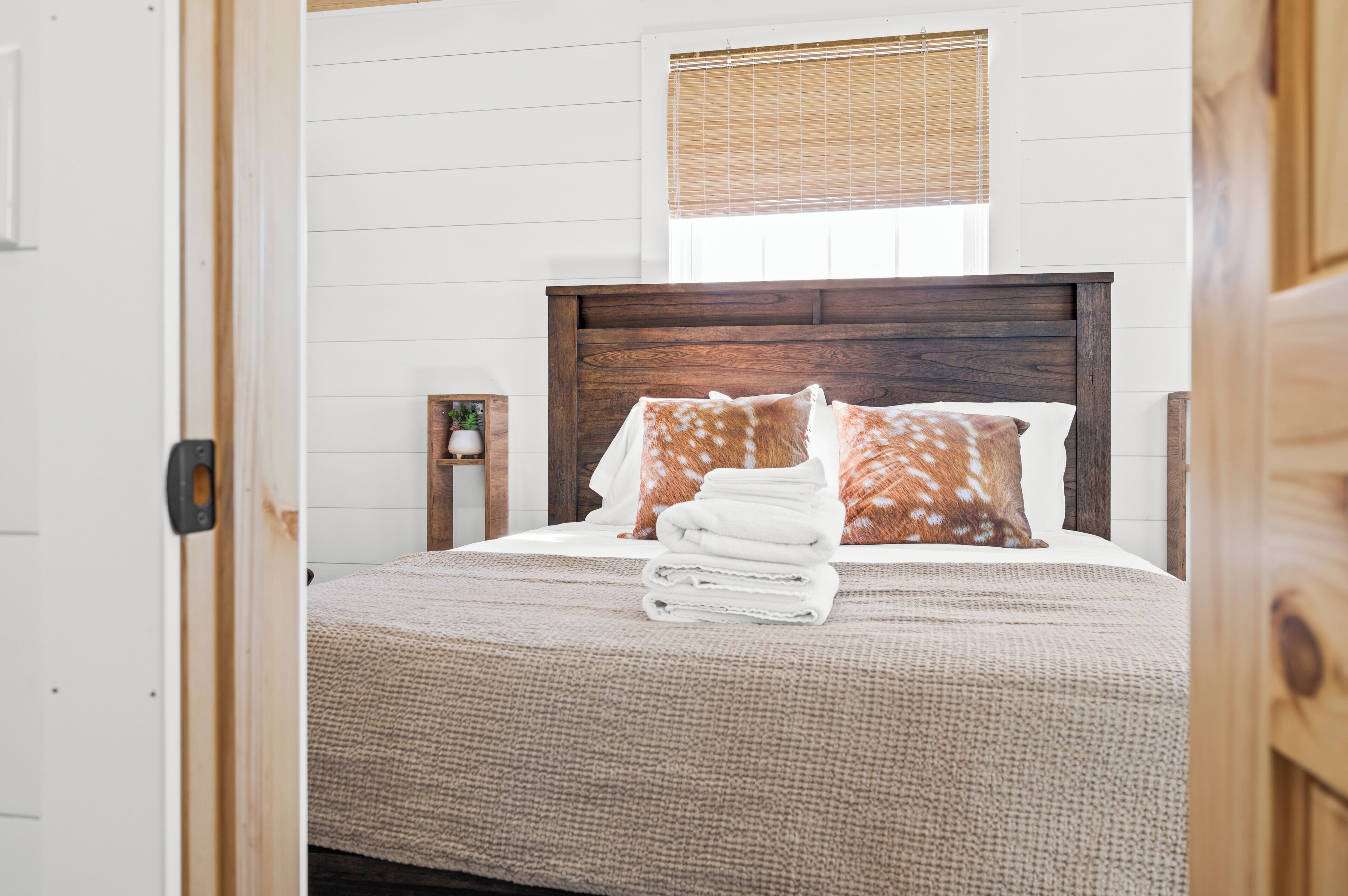 A neatly made bed with a wooden headboard, a window with blinds, and a small bedside table with a lamp.