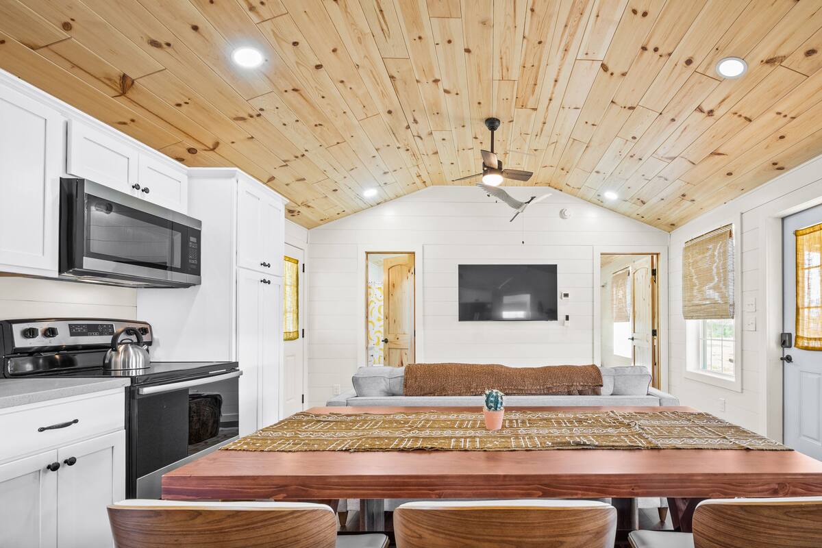 A kitchen with white cabinets, a wooden ceiling, a large wooden dining table, and a TV mounted on the wall.