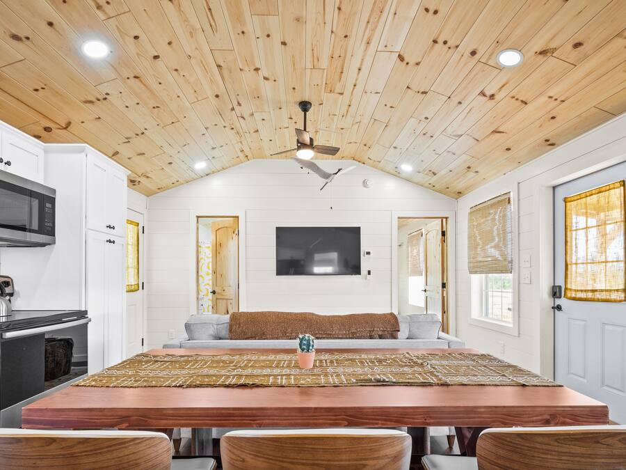 Una cocina con armarios blancos, techo de madera, una gran mesa de comedor de madera y un televisor montado en la pared.