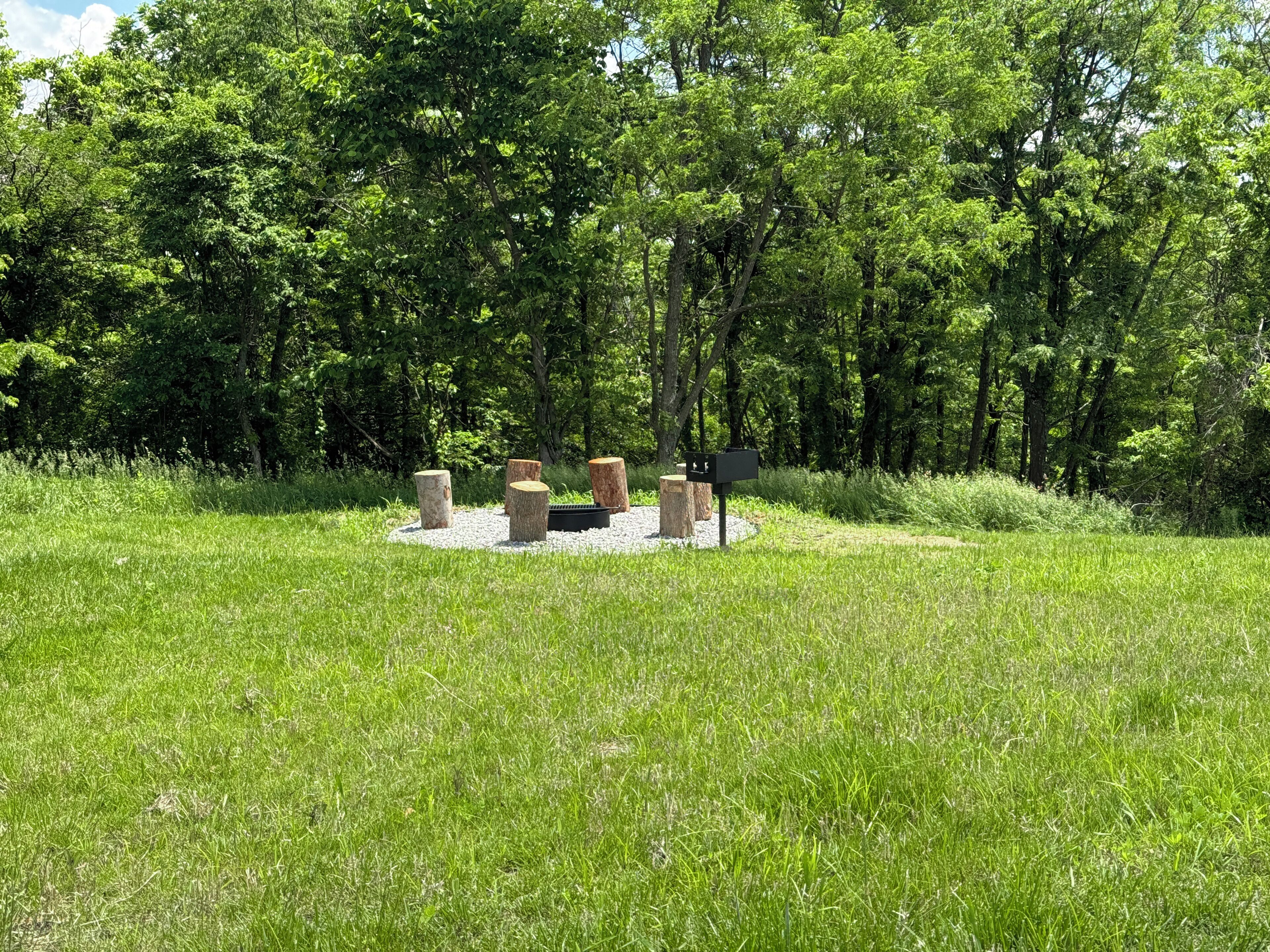 Un espacio de estar al aire libre con forma circular, que incluye un brasero, rodeado de árboles y césped.