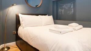 A neatly made bed with white linens and towels, a round mirror above it, and a wall-mounted artwork depicting a horse and a dog. - Cosy 1-bedroom Apartment - Putney High Street (London)
