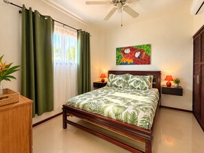 A bedroom with a bed, green curtains, a ceiling fan, a wall-mounted painting, bedside lamps, and a small plant. - Pura Vida Dreams (Tamarindo)