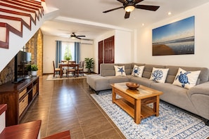 A modern living room with a sofa, coffee table, and TV. There is a staircase with wooden railings and a dining area with a table and chairs. - Pura Vida Dreams (Tamarindo)