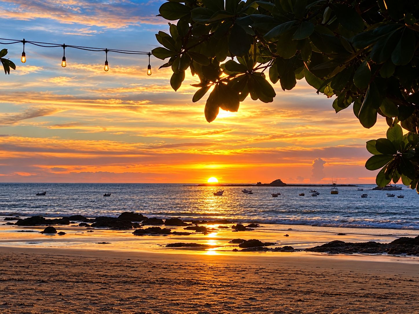 A sunset over a beach with boats and a string of lights.