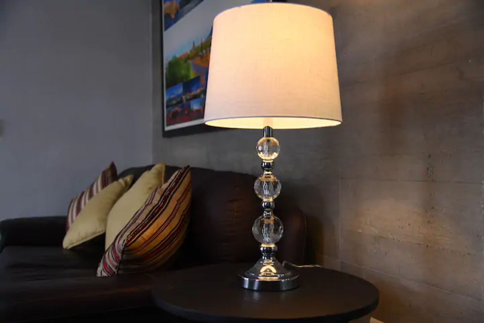 A lamp with a clear glass shade on a round table, a dark-colored sofa with patterned cushions, and a framed picture on the wall.