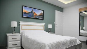 Un dormitorio con cama, mesita de noche, cómoda, televisor montado en la pared y una puerta.