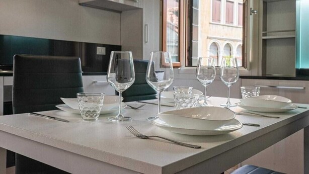 Una mesa de comedor dispuesta con platos, vasos y cubiertos en un interior moderno con vistas a edificios.