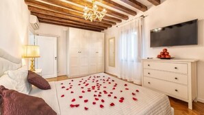 Un dormitorio con techo de madera, cama con pétalos de rosa, televisor y cómoda blanca.
