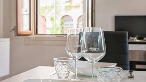 A dining table with two wine glasses and two small glasses, set against a window with a view of buildings. - Casanova Fenice III (Venezia)