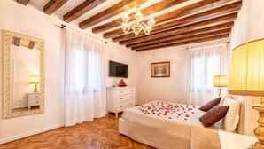 A bedroom with a wooden floor, a bed with rose petals, a mirror, a TV, and a chandelier. - Casanova Fenice III (Venezia)