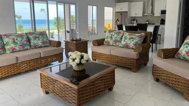A spacious living room with wicker furniture, a coffee table with a vase of flowers, and large windows offering a view of the ocean. - Luxury Beach Front Villa with Private pool and stunning Ocean View! (Punta Rucia)