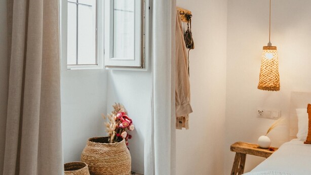Une chambre à coucher avec un lit, une table de chevet avec une lampe, une fenêtre avec des rideaux et deux paniers en osier posés sur une étagère.