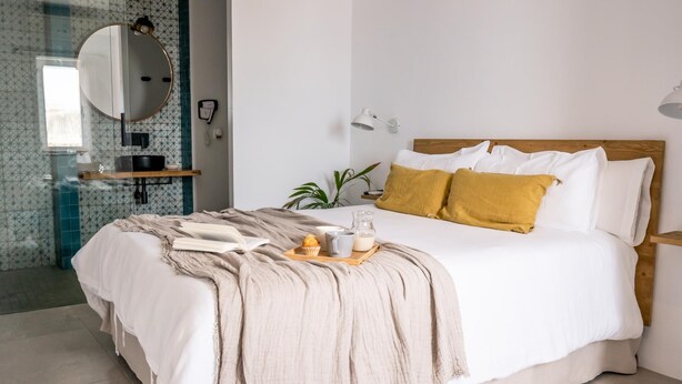 A modern bedroom with a large bed, a bedside table, a mirror, and a bathroom visible through a glass partition.
