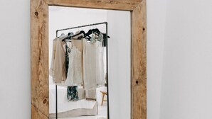 Un miroir à cadre en bois qui reflète un portant à vêtements sur lequel sont disposés différents articles d’habillement.