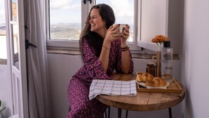 A person sitting by a window with a view of the countryside, holding a cup. There's a table with croissants and a bottle of perfume.