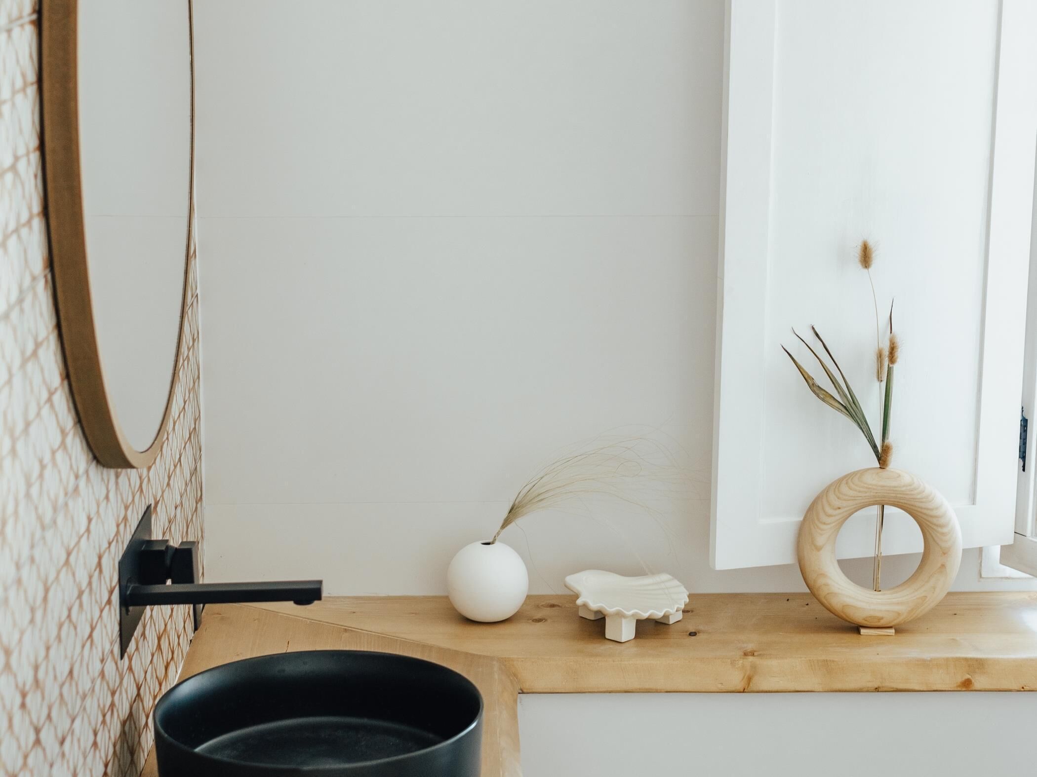 Una cocina con una olla negra sobre la encimera, un jarrón blanco con plantas secas y un anillo de madera en un estante.