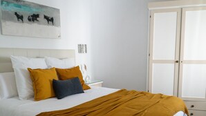 Una cama bien hecha con ropa de cama blanca y color mostaza, una mesita de noche con lámpara, y una puerta con paneles de vidrio esmerilado.