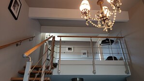Un escalier avec une rampe en bois et des balustrades en métal, menant à un palier du deuxième étage où se trouvent un lustre et un porte-manteau.