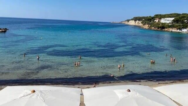 Une plage avec une eau d’un bleu limpide, des gens qui se baignent et prennent des bains de soleil, et des parasols blancs.