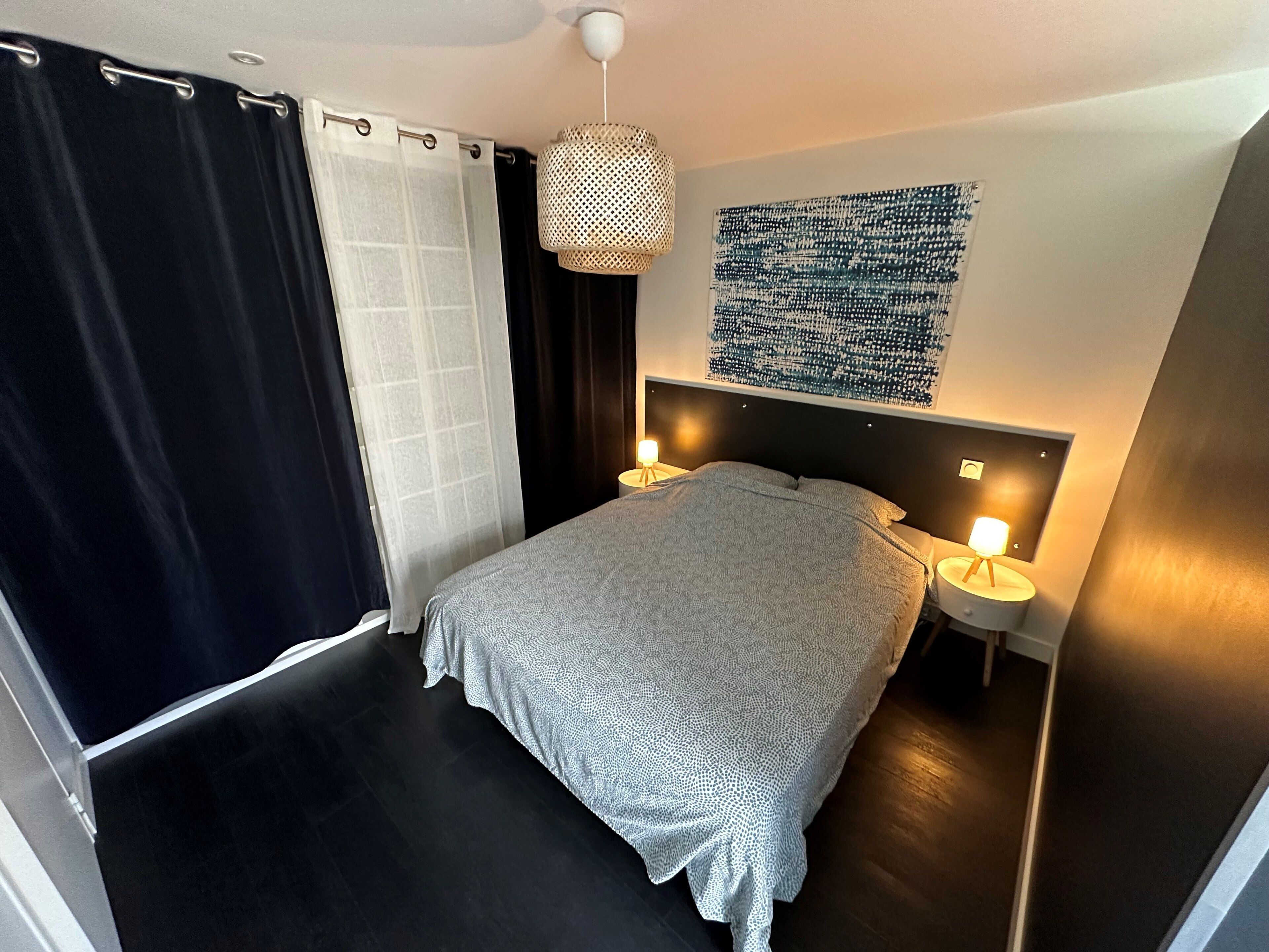 A modern bedroom with a large bed, two bedside lamps, a hanging light fixture, and a wall art piece.