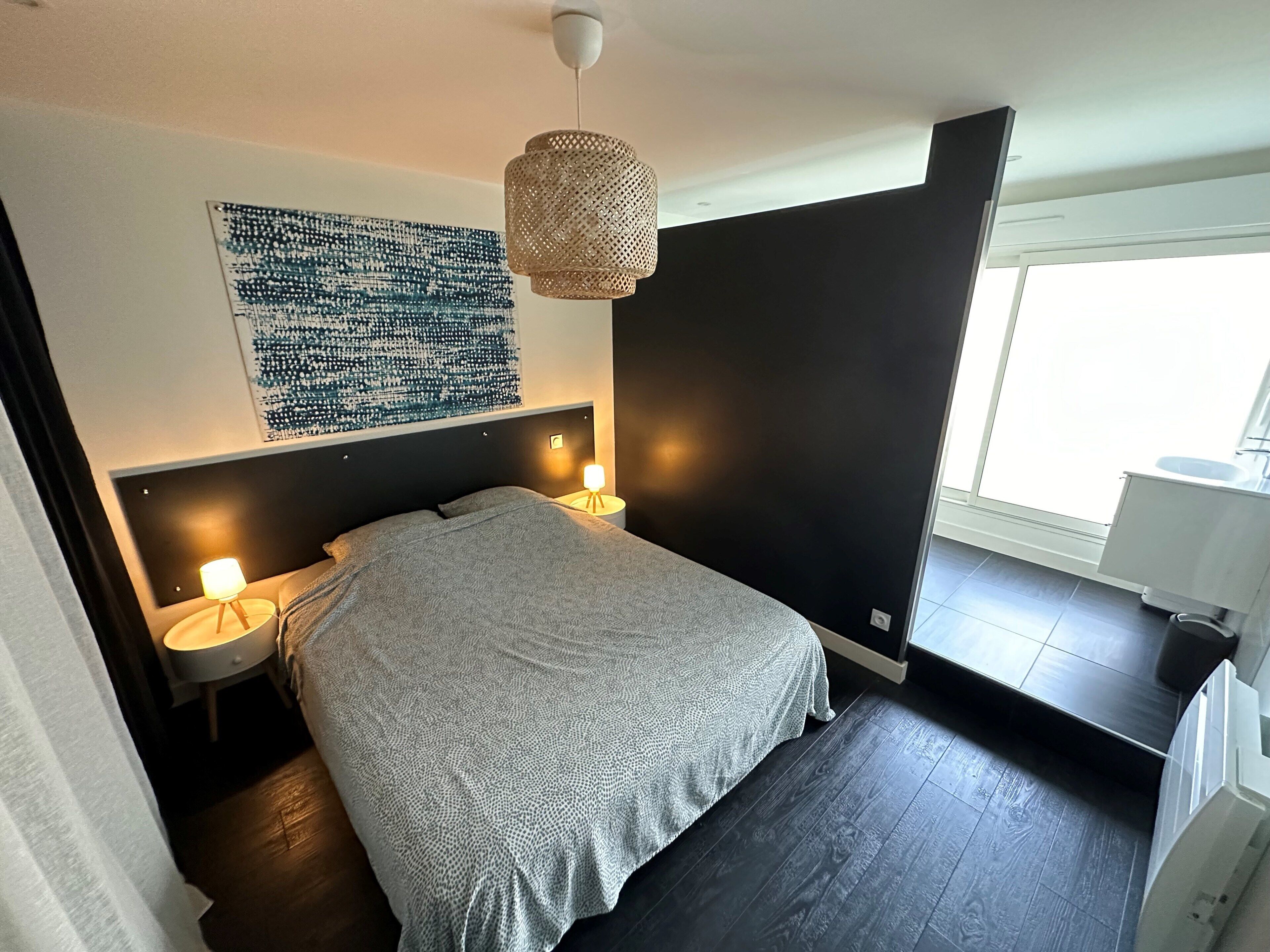 A modern bedroom with a large bed, two bedside lamps, a wall-mounted artwork, and a window with a view.
